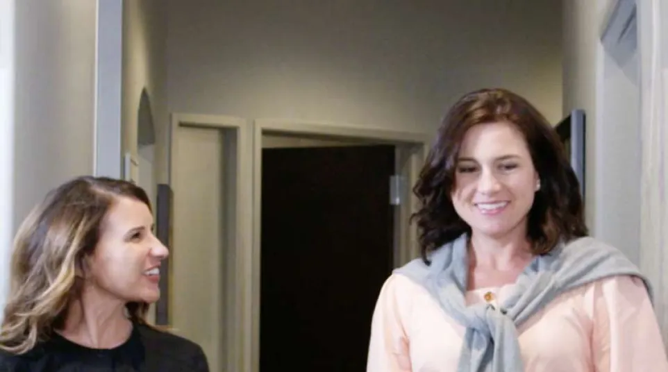 Two women walking and smiling in a hallway, one holding a clipboard, dressed casually and professionally.