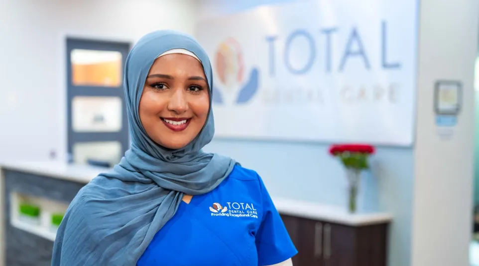 Smiling female dental professional in blue scrubs and hijab standing inside Total Dental Care clinic.