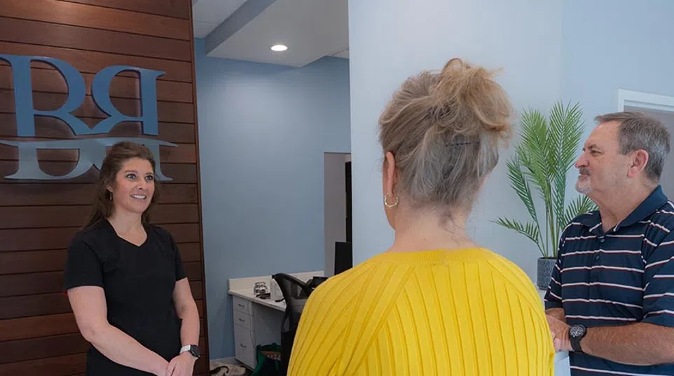 Receptionist in black talking to two visitors at a modern office front desk with wooden accent wall.