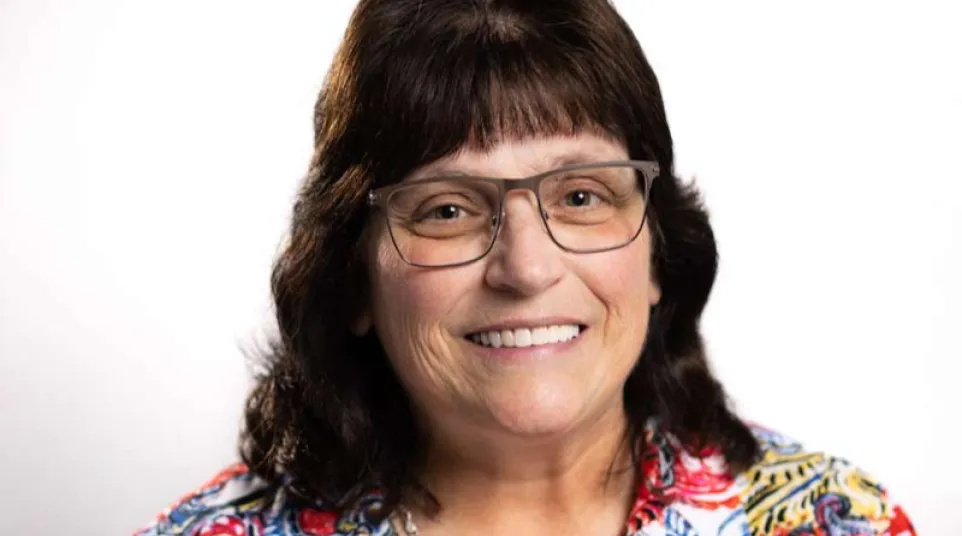 Woman with glasses smiling brightly wearing a colorful patterned shirt against a white background.