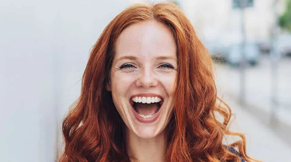 Young woman with long curly red hair smiling joyfully outdoors in casual dark blue top
