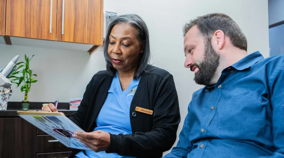 Female doctor in blue scrubs explaining medical information to male patient in clinic office.