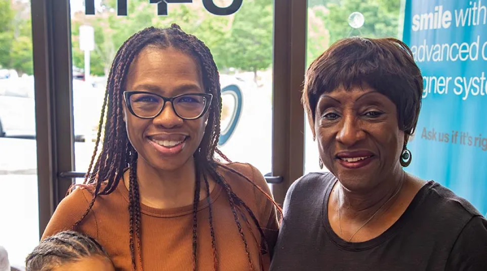 Three generations of smiling African American women standing indoors near a window with a sign in the background.