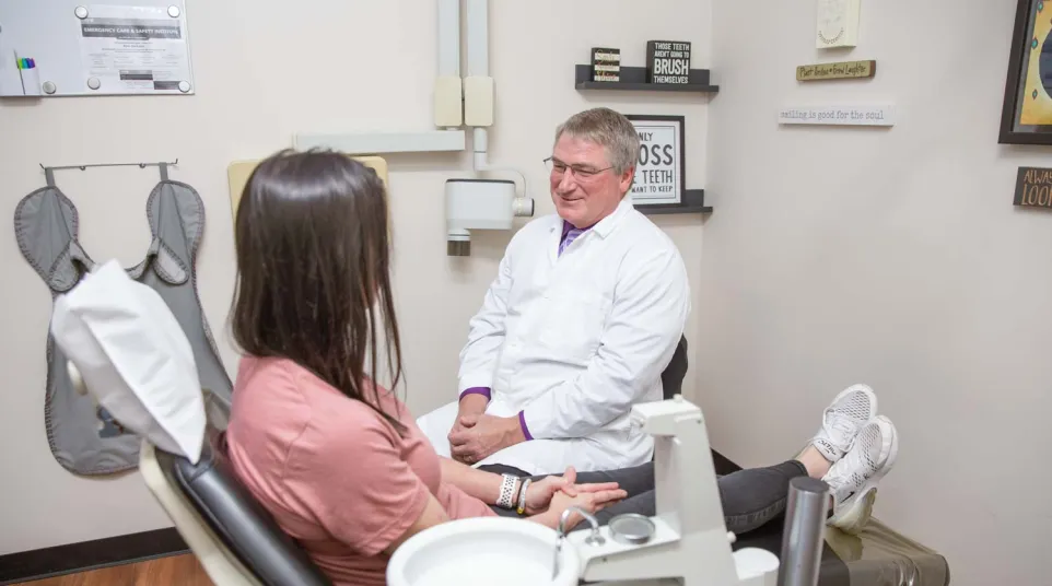 Dentist in white coat talking to a female patient sitting in a dental chair in a clinic room.