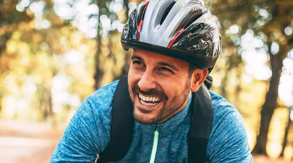 Smiling man in blue jacket and helmet leaning on bicycle handlebars in autumn forest.