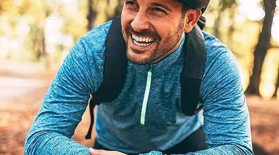 Smiling man in a blue jacket and helmet leaning on his bicycle handlebars in a forest with autumn leaves.