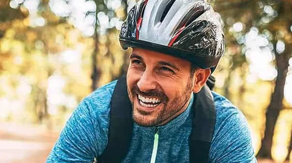 Smiling man in a blue jacket and helmet leaning on his bicycle handlebars in a forest with autumn leaves.