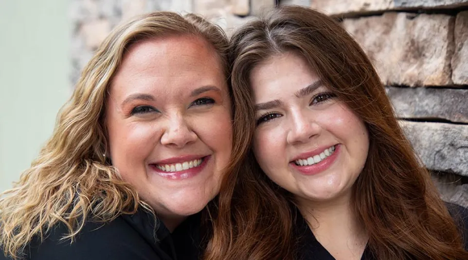 Two women wearing black scrubs smiling and embracing in front of a rustic stone wall background.