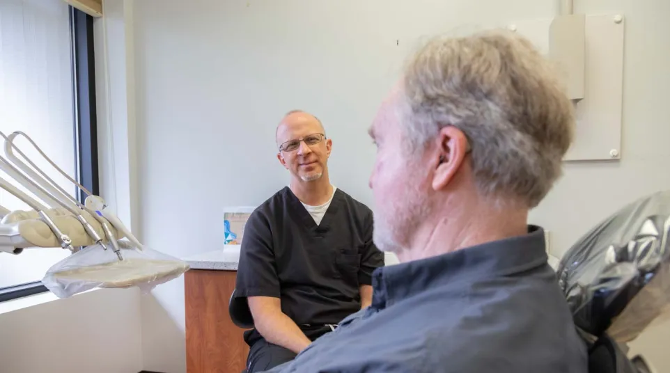 Dentist in black scrubs consulting with male patient in dental office with natural light