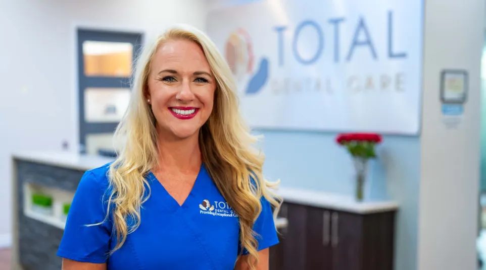 Smiling female dental professional in blue scrubs standing inside Total Dental Care clinic with logo in background.