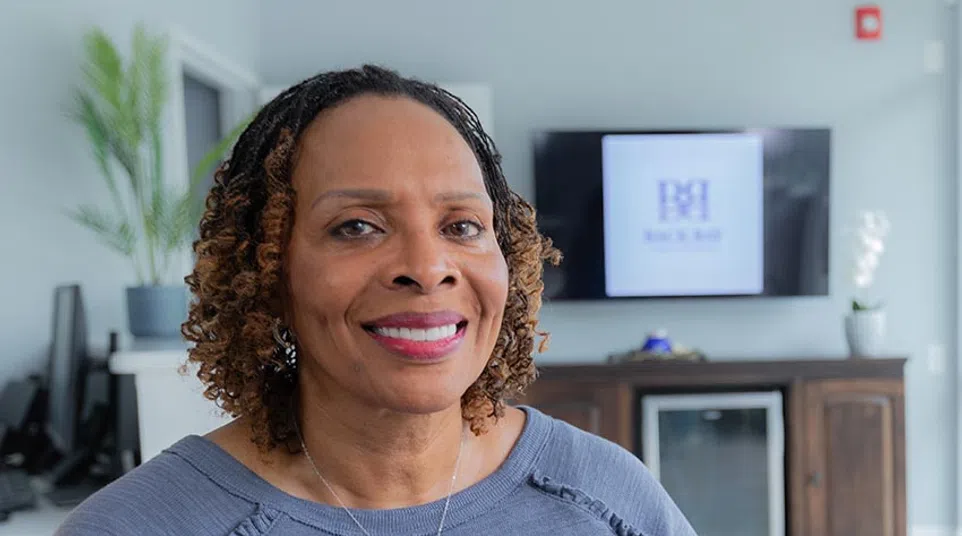 Smiling woman with curly hair wearing a blue top in a modern office setting with a TV and plants in background