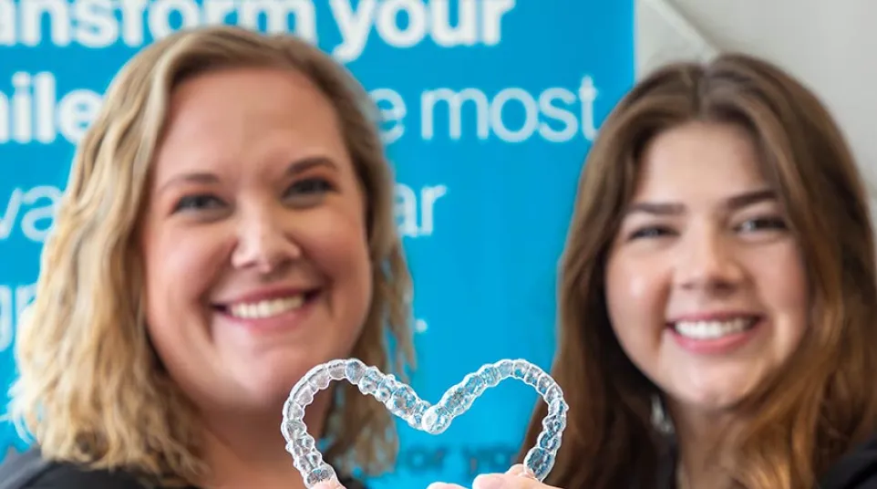 Two smiling women holding a clear dental aligner shaped like a heart in a dental office.