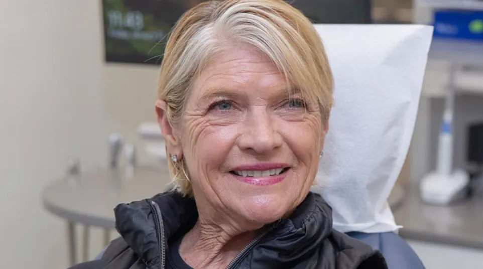 Smiling elderly woman with short blonde hair wearing a black vest sitting in a medical or dental office chair.