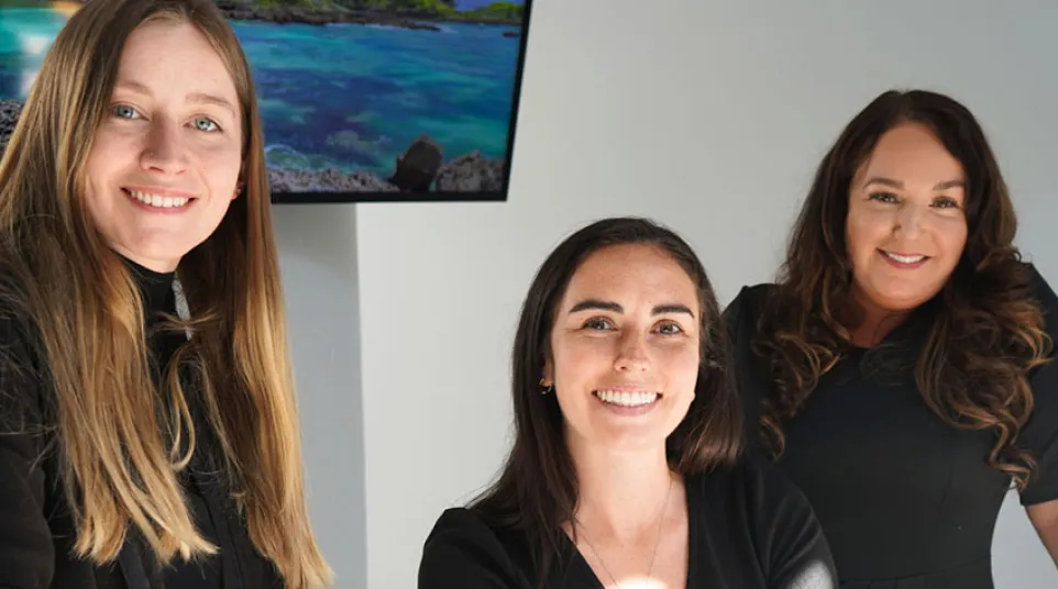 Three professional women smiling in an office setting with a ocean landscape screen in the background