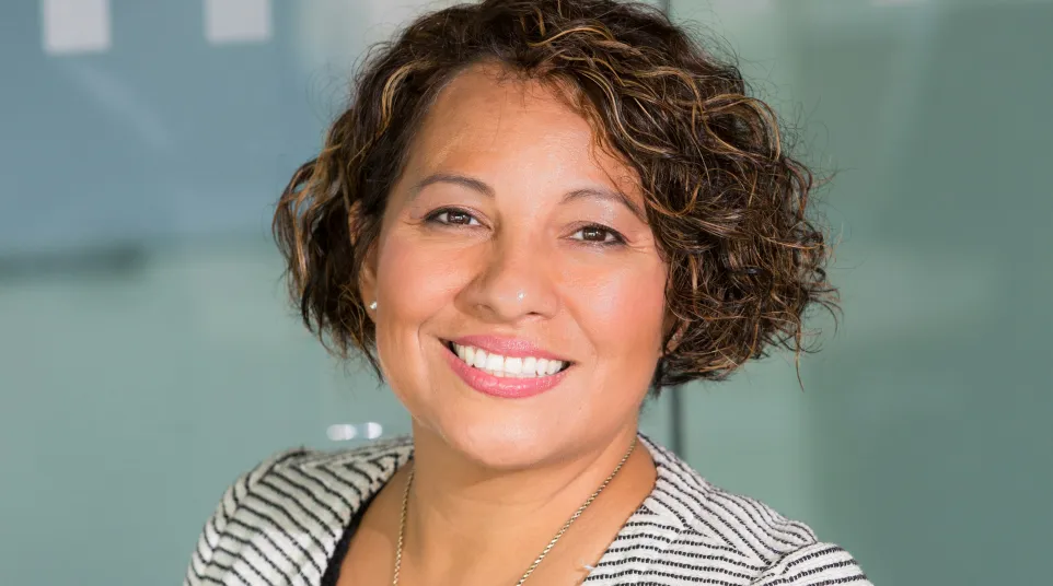 Smiling confident woman with curly hair wearing a black top and striped blazer in an office setting.