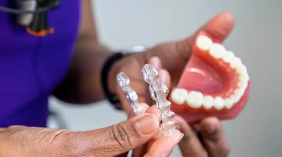 Smiling young woman inserting clear dental aligners over her teeth indoors with warm lights in the background