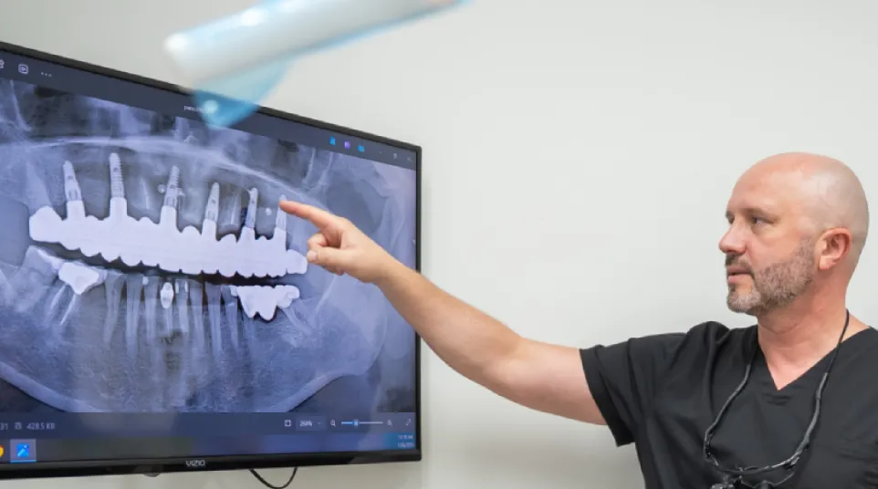 Dentist pointing at dental X-ray displayed on monitor showing teeth and implants in a clinical setting.