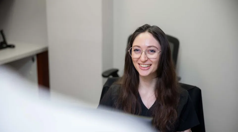 Smiling woman with long brown hair and glasses sitting in an office chair in a modern workspace.