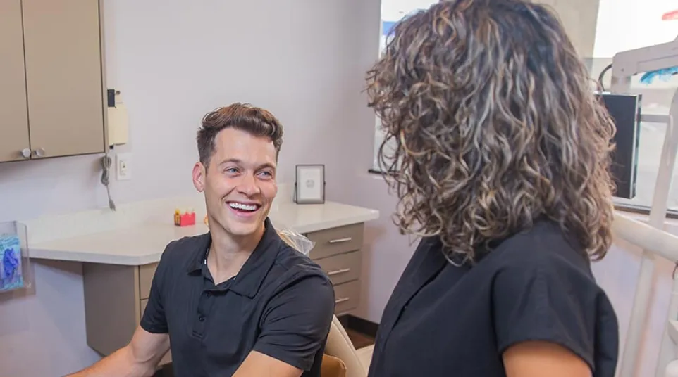 Smiling man sitting at a desk with a computer talks to a standing woman in a casual office setting.
