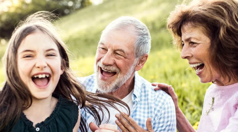 Happy grandparents laughing and playing outdoors with their granddaughter in a sunny green meadow.