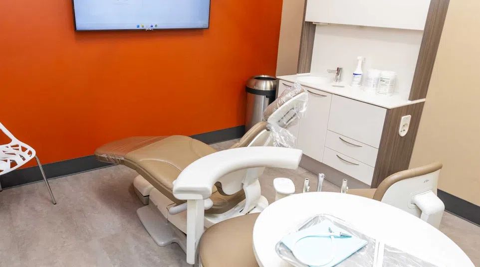 Modern dental office with beige chair, orange accent wall, white shelving, and screen displaying Renew Dental Arts logo.
