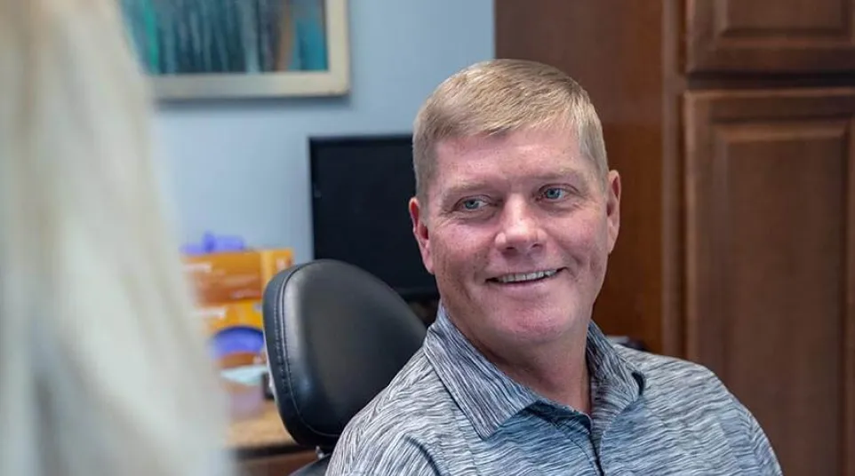 Man in gray polo shirt smiling while sitting in office chair talking to a person in blue attire.