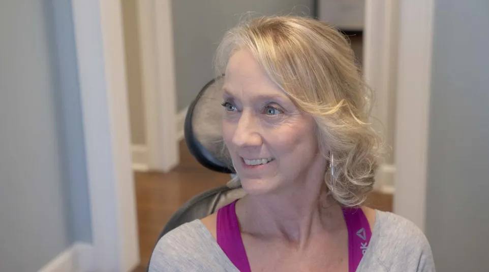 Smiling middle-aged woman with blonde hair wearing casual workout clothes sitting in a chair indoors.