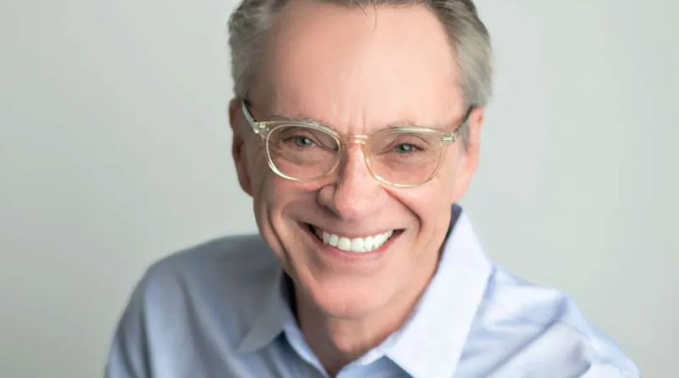 Smiling middle-aged man wearing glasses and a light blue button-up shirt against a plain gray background