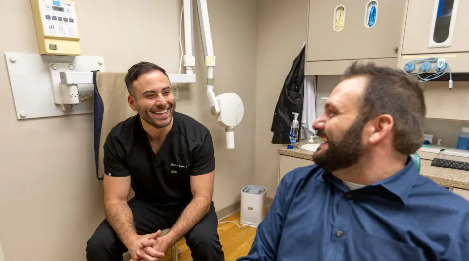Smiling dentist in black scrubs converses with male patient in a clinic room with dental equipment.