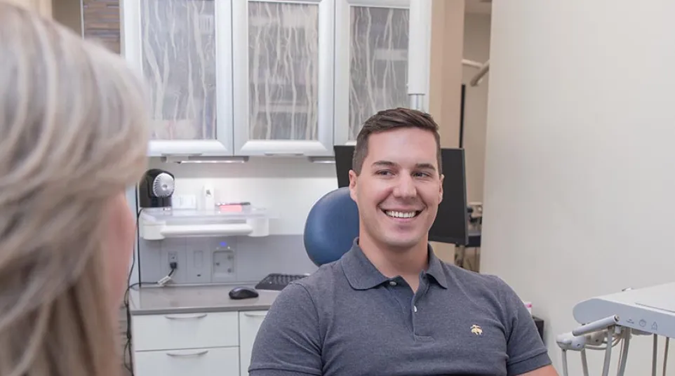 Smiling man sitting in a dental chair talking to a female dentist in a modern clinic room.