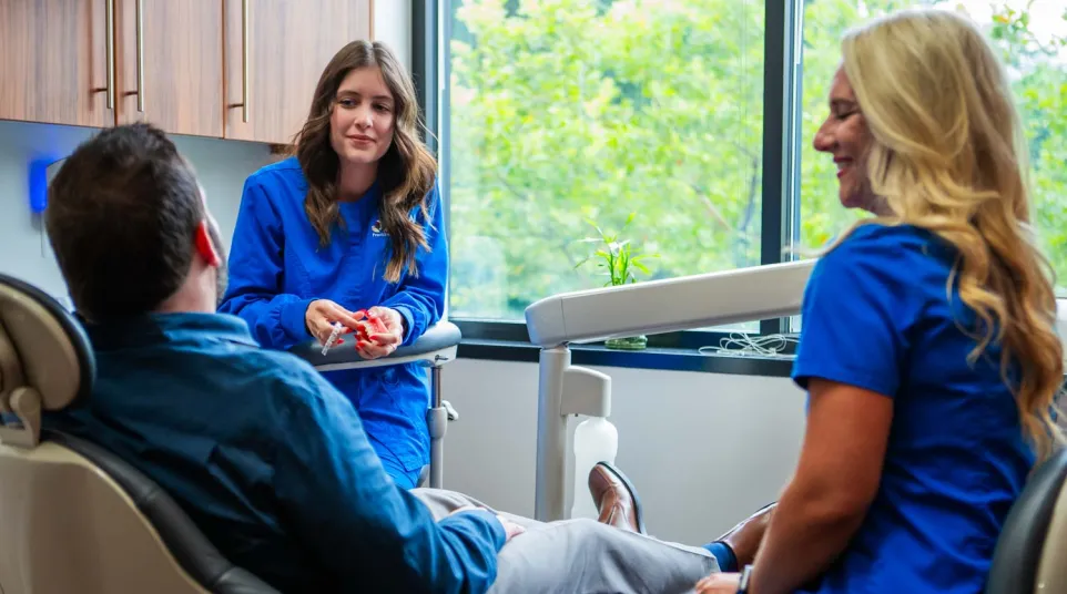 Dental hygienists in blue scrubs consulting a male patient reclining in a dental chair near a large window.