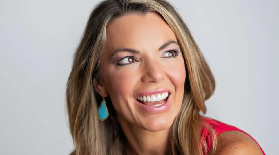 Smiling woman with long blonde hair wearing a red top and turquoise earrings looking to the side.