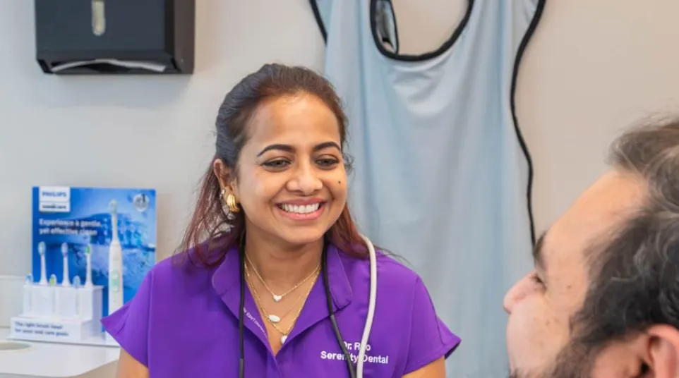 Smiling female dentist in purple scrubs talking with male patient in dental office setting.
