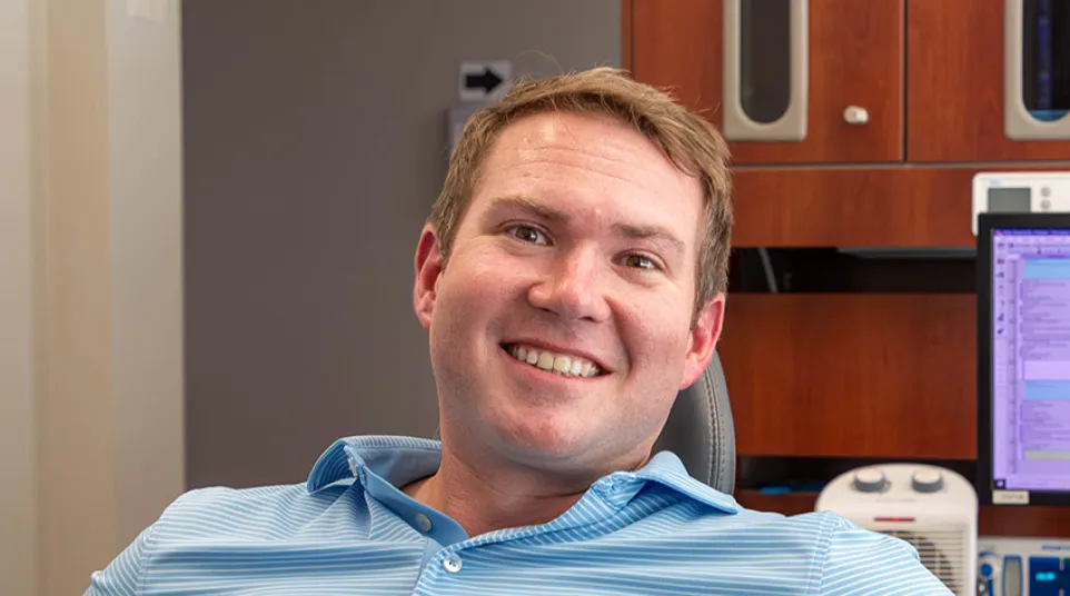 Smiling man in a light blue striped polo shirt sitting in a chair in an office setting with computer and wooden cabinets.