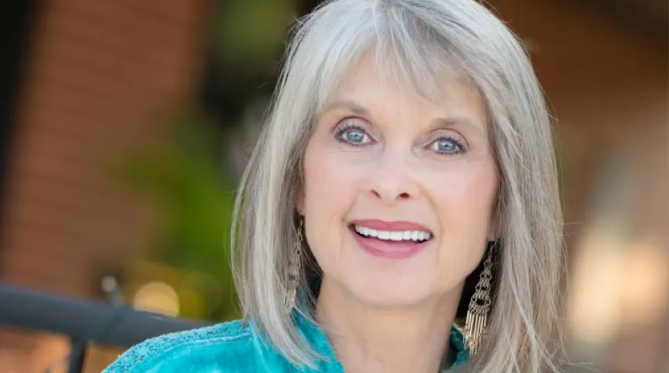 Smiling elderly woman with gray hair wearing a turquoise embroidered blouse and dangling earrings outdoors.