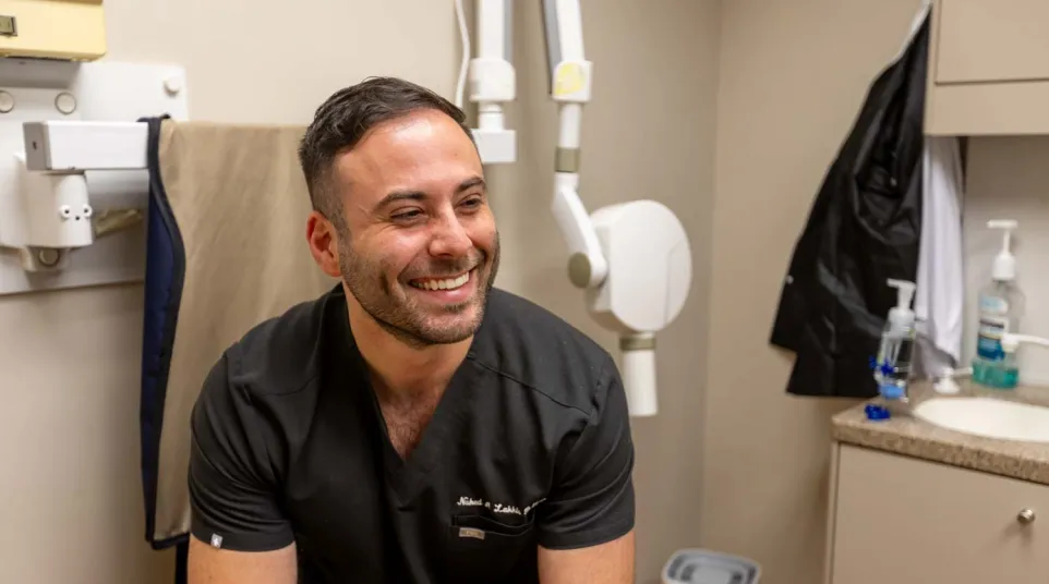 Smiling male dentist in black scrubs sitting in dental office with equipment and sink in the background