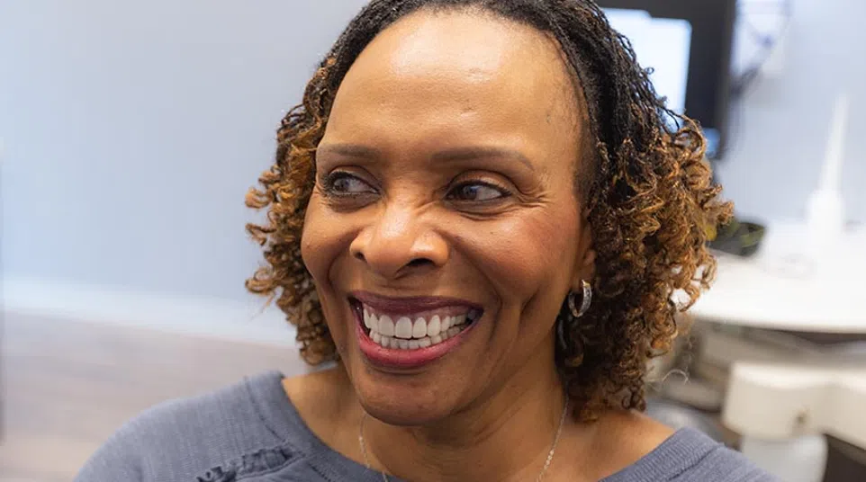 Smiling woman with curly hair and silver necklace sitting indoors wearing a gray top