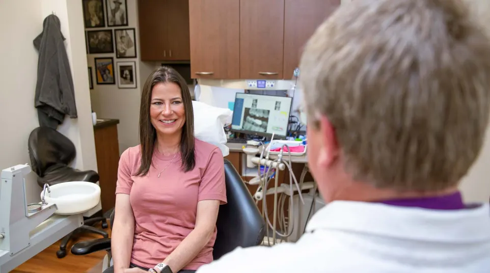 Smiling woman in pink shirt sitting in dental office talking to dentist with dental equipment in background