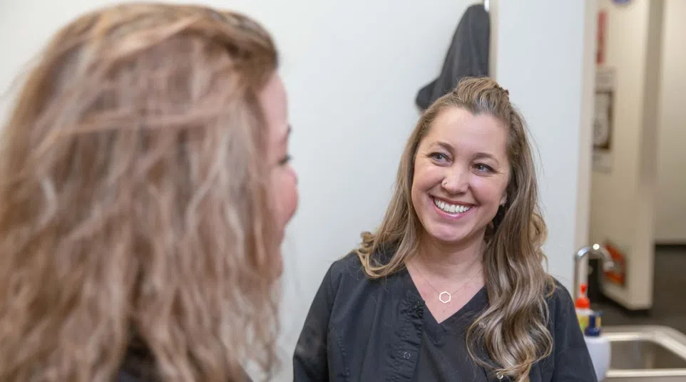 Smiling woman with long hair wearing black scrubs standing in a bright room with a sink and hand sanitizer.