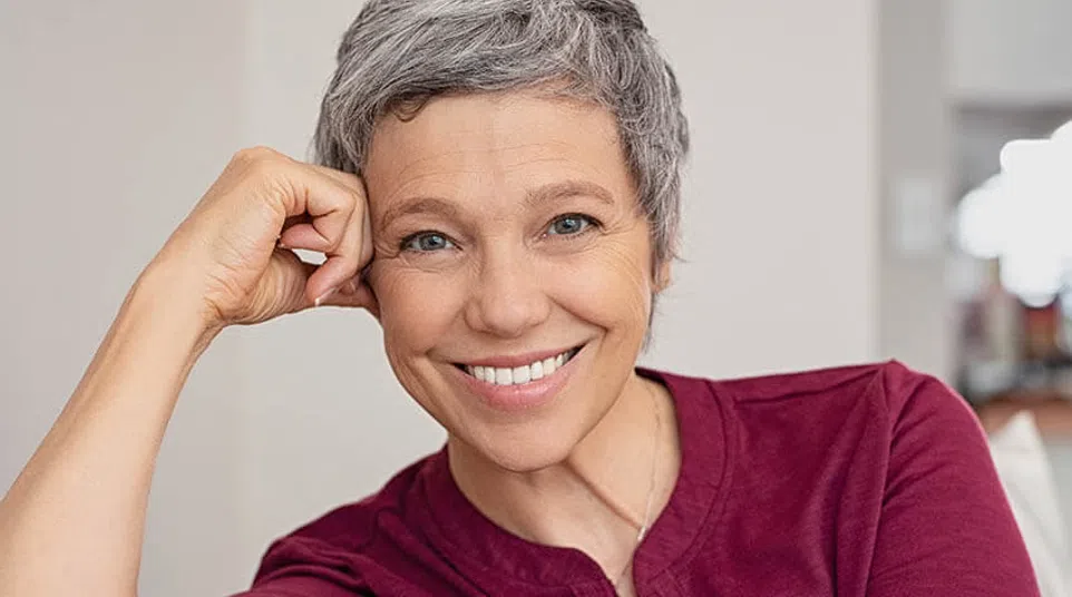 Smiling woman with gray hair in a maroon shirt, sitting comfortably and resting her chin on her hand.