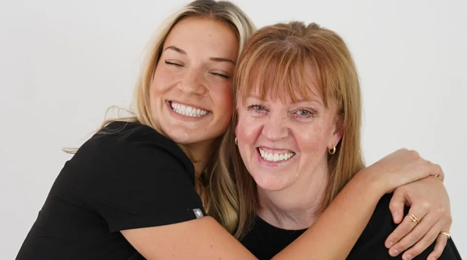 Two women wearing black scrubs smiling and hugging against a plain white background, showing warmth and friendship.