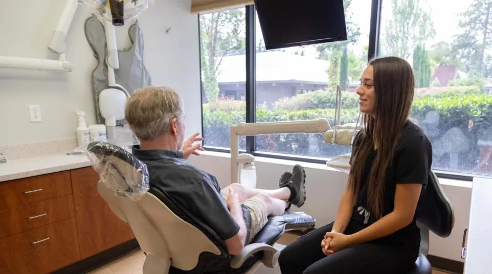 Dentist consulting an elderly male patient in a bright dental office with large windows and modern equipment.