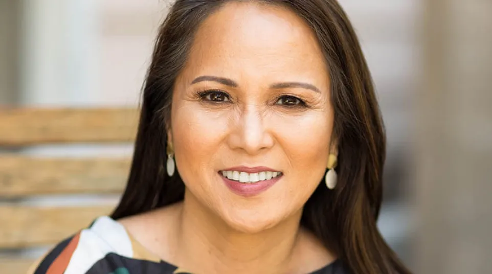 Smiling middle-aged woman with long dark hair wearing colorful patterned top and white earrings outdoors