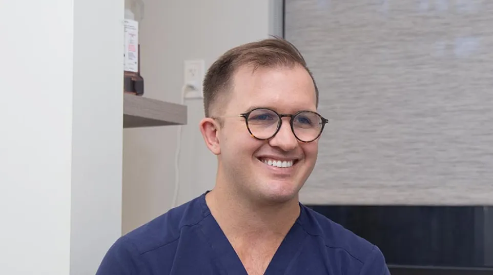 Smiling man in navy scrubs and glasses sitting indoors in a medical or clinical setting.