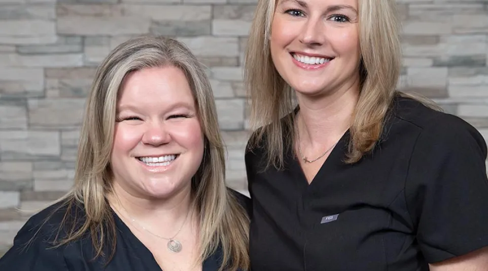 Two smiling women in black medical scrubs standing in front of a stone wall background.