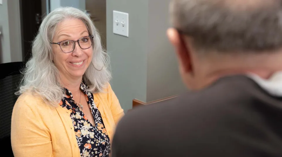 Smiling woman with gray hair and glasses wearing a yellow cardigan talks to a man in an office setting.