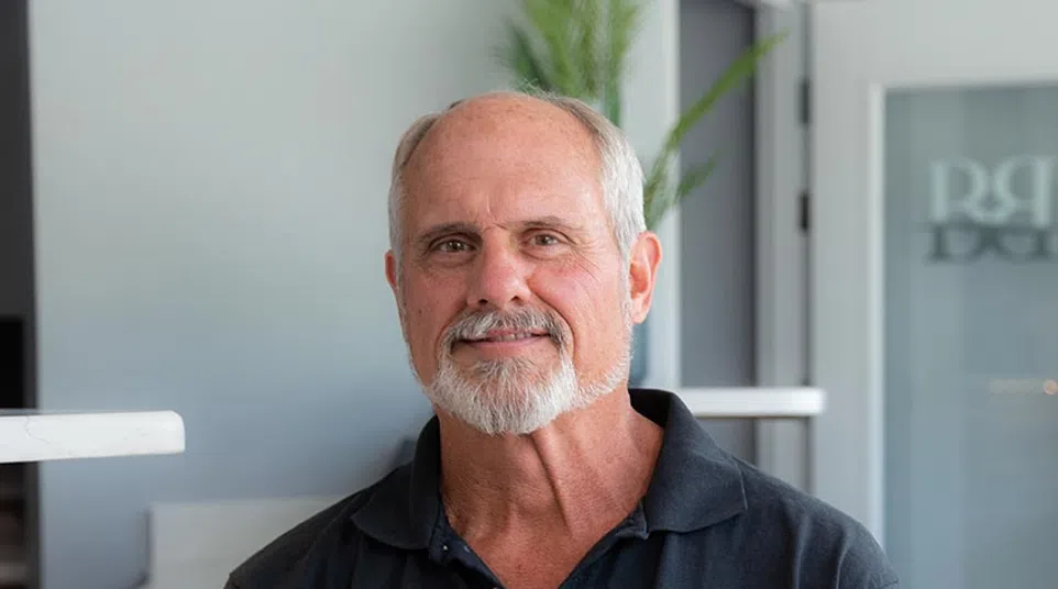 Smiling older man with gray beard wearing a black Strayhart Plumbing polo shirt sitting indoors.