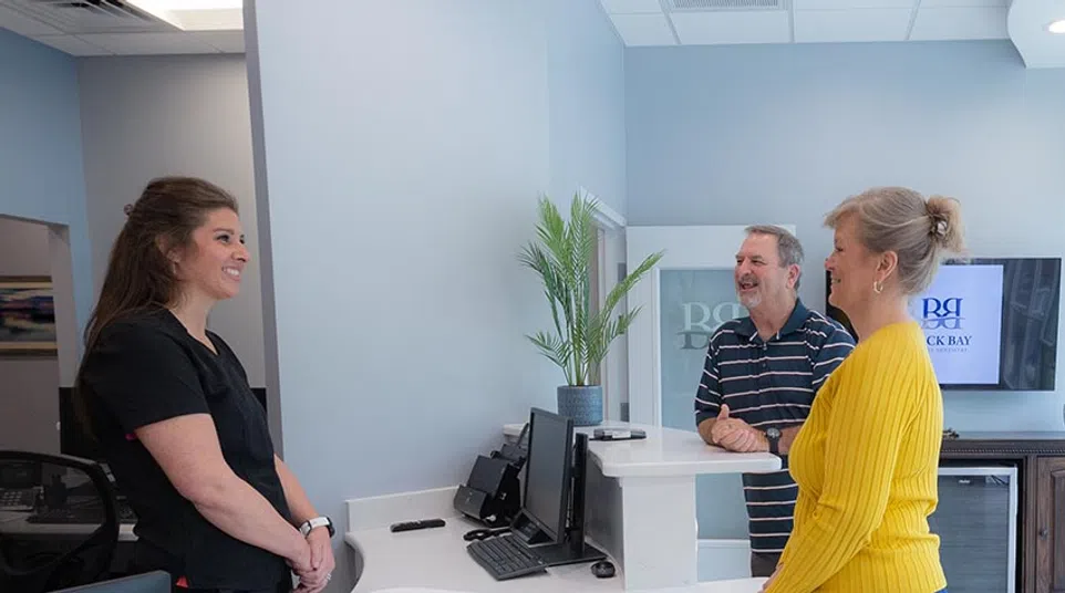 Receptionist talking to an elderly couple at a modern medical office with a white marble counter and indoor plant.