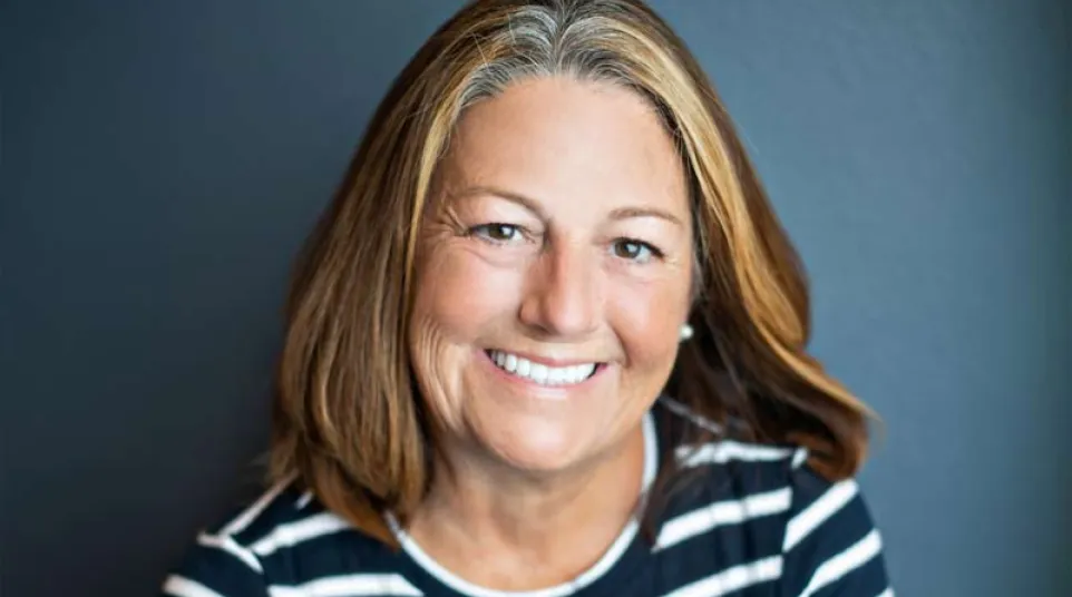 Smiling middle-aged woman with blonde hair wearing a black and white striped shirt against a dark background.
