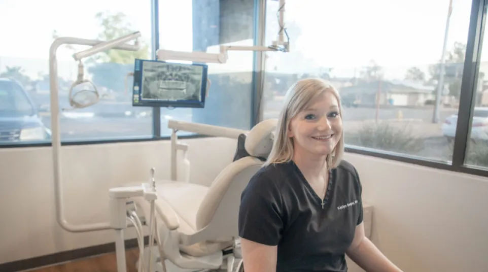 Smiling female dentist in black scrubs sitting at desk in a modern dental office with dental chair and equipment.